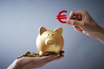 Une main s'apprête à glisser un symbole euro dans une tirelire cochon posée sur une autre main pleine de pièces de monnaie