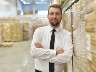 Dans un entrepôt, un homme souriant, portant chemise blanche et cravate, se tient appuyé, bras croisés, contre un chargement de cartons