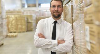 Dans un entrepôt, un homme souriant, portant chemise blanche et cravate, se tient appuyé, bras croisés, contre un chargement de cartons